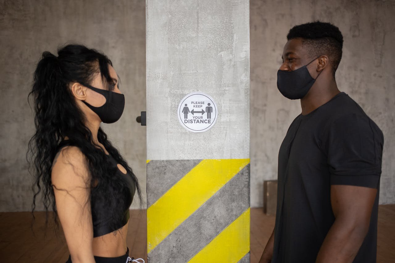 Two people practicing social distancing with masks indoors near a safety sign.