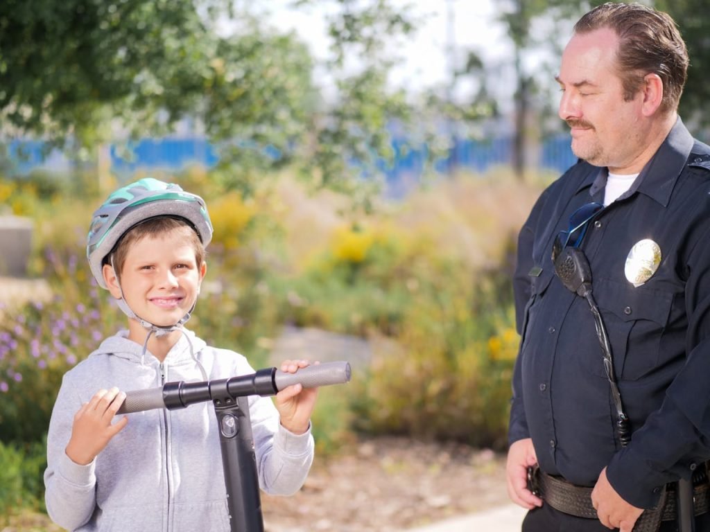 Mastering the First Impression: Your intriguing post title goes here A police officer smiling at a boy on an electric scooter in a park setting, promoting community safety.